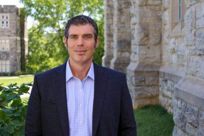 portrait of man in shirt and blazer outside next to stone building